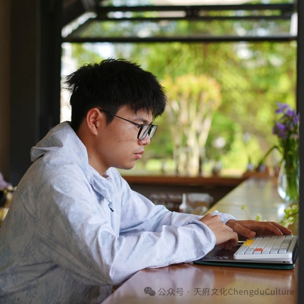 Leo Wang wearing glasses working in a serene environment, with green plants outside the window in the background.