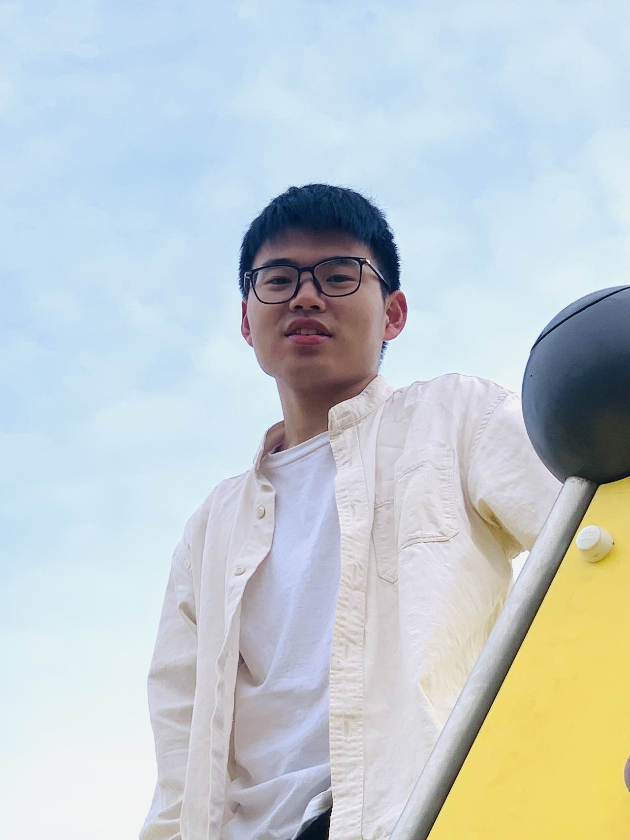 Leo Wang wearing glasses, dressed in a white shirt and white t-shirt, standing next to a yellow structure with a clear sky in the background.